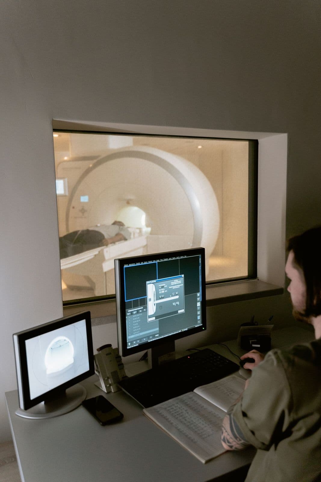 Técnico acompanhando um exame de tomografia pela sala de comando, com o paciente visível através do vidro